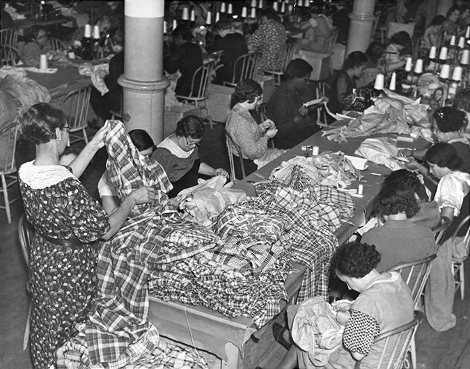 Costume workshop with long sewing tables where seamstresses cut and stitch plaid fabric for stage costumes